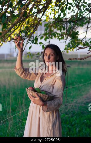 Schönes Mädchen im Apfelgarten. Eine junge Frau in einem schönen hellen Kleid sammelt grüne Äpfel in einem Korb zwischen Apfelbäumen. Ernte A Stockfoto