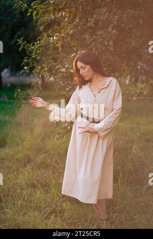 Schönes Mädchen im Apfelgarten. Eine junge Frau in einem schönen hellen Kleid sammelt grüne Äpfel in einem Korb zwischen Apfelbäumen. Ernte A Stockfoto