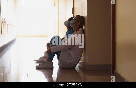 Nahaufnahme von erschöpften Schwestern nach harter langer Operation auf dem Boden in der Halle sitzend, schwierige Schicht. Medizin, Leben retten, Gesundheitswesen, Klinik, Hilfekonzept Stockfoto