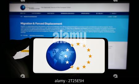 Person, die ein Mobiltelefon mit dem Logo des Europäischen Auswärtigen Dienstes (EAD) vor der Webseite hält. Schwerpunkt auf dem Display des Telefons. Stockfoto