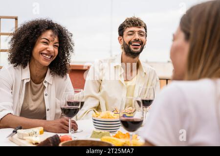 Abwechslungsreiche junge Leute, die an einem Terrassentisch sitzen und Spaß haben. Fröhliche Gruppe von Freunden, die lachen und plaudern, während sie draußen Drinks trinken. Stockfoto