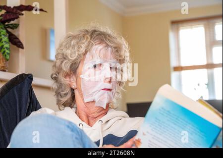 Reife Frau, die zu Hause ein Buch liest, trägt eine Gesichtspackung, um unerwünschte Falten loszuwerden und ihre Haut glatt zu halten Stockfoto