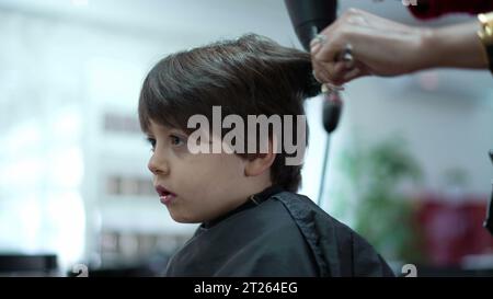 Little Boy wird vom professionellen Friseur im Friseursalon geschnitten und gekämmt, Person mit Haartrockner Stockfoto