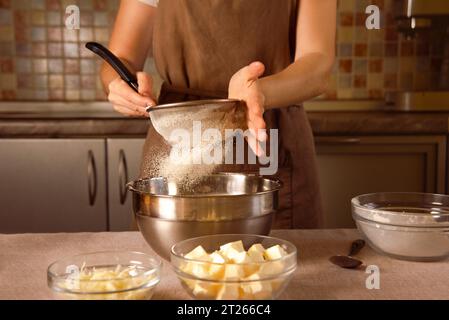 Die Küchenchefin in brauner Schürze siebt Mehl durch Sieb für die Herstellung von Teig auf Küchenhintergrund Stockfoto