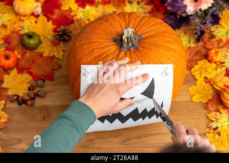 Augsburg, Bayern, Deutschland. Oktober 2023. Eine Halloween-Laterne vorbereiten, einen Kürbis aushöhlen und schnitzen. Mann, der zu Hause in einem Halloween-Kürbis mit Schablone und Messer ein böses Gesicht schnitzt *** Vorbereitung einer Halloween-Laterne, Aushöhlen und Schnitzen von einem Kürbis. Mann schneidet ein fieses Gesicht Zuhause in einem Halloweenkürbis mit einer Schablone und einem Messer Credit: Imago/Alamy Live News Stockfoto