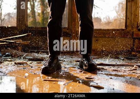 Männliche Beine in schwarzen Hosen und Turnschuhen zwischen der Pfütze und gefrorenen Tropfen in einem überfluteten, zerstörten und verlassenen Haus. Sonnenstrahlen. Stockfoto