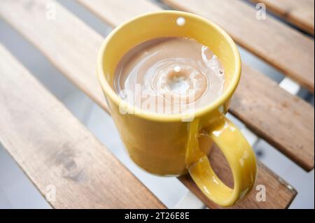 Ein Tropfen Milch, der auf eine Tasse Kaffee fällt und Milch auf einen Holztisch auf einer Terrasse gestellt wird Stockfoto