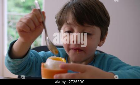 Kind isst weich gekochtes Ei mit Löffel, kleiner Junge knackt die Spitze von Oeuf à la Coque, isst cremigen Protein-Snack Stockfoto