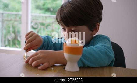 Kind isst weich gekochtes Ei mit Löffel, kleiner Junge knackt die Spitze von Oeuf à la Coque, isst cremigen Protein-Snack Stockfoto