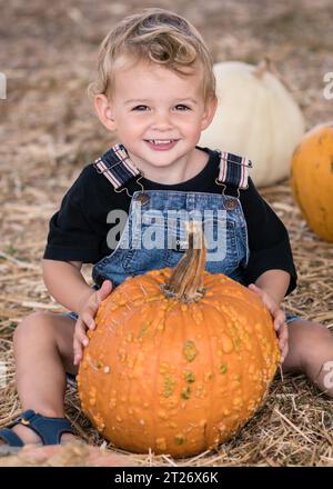 Porträt eines 2-jährigen Jungen, der lächelt und einen orangefarbenen Kürbis in der Hand hält, auf der Deluca Farm in San Pedro, CA. Stockfoto