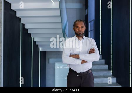 Porträt eines afroamerikanischen Mannes in einem weißen Hemd, der in einem Bürozentrum steht und seine Arme auf der Brust kreuzt, selbstbewusst und ernsthaft in die Kamera schaut. Stockfoto