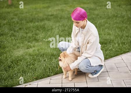Stilvolle Frau mit lila Haaren, die süße pommersche Spitz auf dem Gehweg im Park in der Nähe von grünem Rasen umhüllt Stockfoto