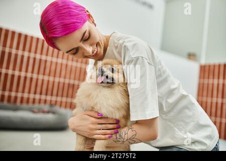 Lächelnde Hundesitterin mit lila Haaren kuschelt süß pommerschen Spitz im Haustierhotel, liebevolle Pflege Stockfoto