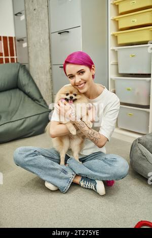 Tätowierter Haustier-Sitter mit lila Haaren, der sich um einen flauschigen Hund schmiegt und in die Kamera auf dem Boden des Haustierhotels lächelt Stockfoto