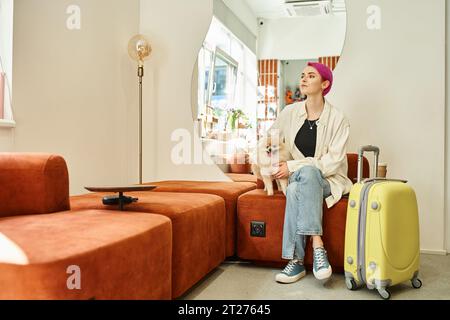 Lila-haarige Frau mit pommerschen Spitz und Koffer wartet in der Lobby des Haustierhotels Stockfoto