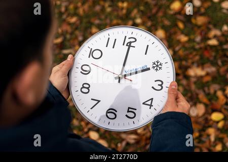 Augsburg, Bayern, Deutschland - 17. Oktober 2023: Mann hält große Uhr mit Winterzeit in der Hand vor Herbstlaub im Freien. Symbolische Änderung der Bildzeit. Text auf Uhr: Winterzeit und Eissymbol FOTOMONTAGE *** Mann hält im freien eine große Uhr mit Winterzeit in der Hand vor herbstlichen Blättern. Symbolbild Zeitumstellung. Text auf Uhr: Winterzeit und Eis Symbol FOTOMONTAGE Credit: Imago/Alamy Live News Stockfoto