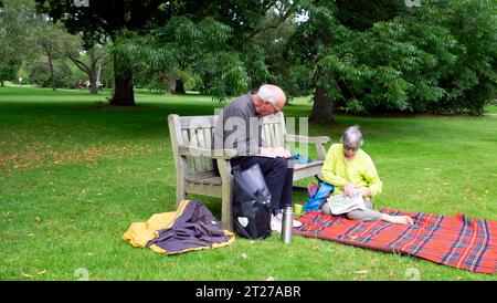 Ältere Ehepaar Mann Frau sitzt auf hölzernen Gartenbank und Picknickdecke, liest Zeitung und trinkt Kaffee Tee Thermos im Sommer Kew Gardens UK Stockfoto