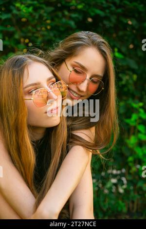 Erstaunte glückliche junge Zwillingsschwestern Generation Zkuschelte sich mit Sonnenbrille in einem Park, Frauen posierten draußen. Konzept der Freundschaft Happy Girls Umarmung Stockfoto