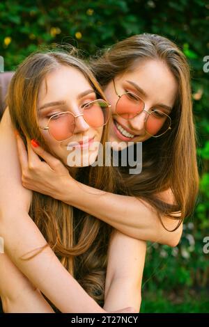 Erstaunte glückliche junge Zwillingsschwestern Generation Zkuschelte sich mit Sonnenbrille in einem Park, Frauen posierten draußen. Konzept der Freundschaft Happy Girls Umarmung Stockfoto