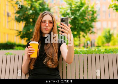 Lächelndes, lustiges Mädchen mit cooler Sonnenbrille, das soziale Medien benutzt und ein Smartphone auf einer Bank in der Stadt sitzt, eine Einwegkaffeetasse aus Papier hält Stockfoto