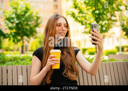 Lächelndes, lustiges Mädchen mit cooler Sonnenbrille, das soziale Medien benutzt und ein Smartphone auf einer Bank in der Stadt sitzt, eine Einwegkaffeetasse aus Papier hält Stockfoto