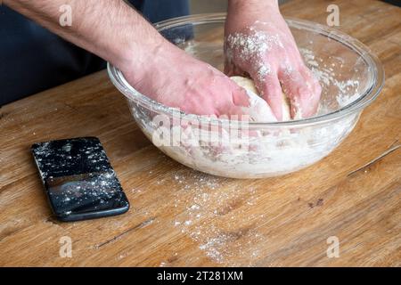 Die Hände des Mannes kneten Teig in einer Glasschale. Während er das Rezept auf einem Handy auf dem Tisch ansieht. Stockfoto