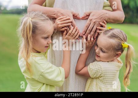 Die Liebe der Schwestern blüht, während sie den schwangeren Bauch ihrer Mutter zärtlich umarmen und ihre Vorfreude und Zuneigung für ihre bald ankommenden Geschwister teilen Stockfoto