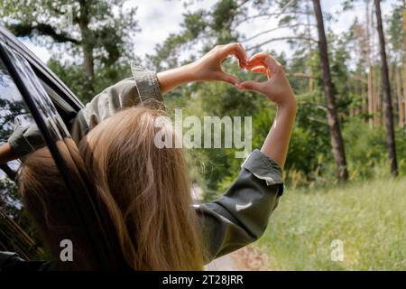 Frau, die im Auto fährt, die Hand aus dem Fenster hält und ein Herzsymbol mit ihren Händen zeigt. Love and Adventure Love Love and Adventure Local Travel Freiheit und Verbindung mit der Natur. Roadtrip mit nachhaltigem Lebensstil am Wochenende zum Entspannen und Ausruhen Stockfoto