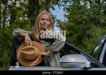 Blonde Frau mit Hut, die neben der Autotür wohnt. Junge Touristen erkunden lokale Reisen und machen echte Momente. Wahre Emotionen Ausdruck des Ausgehens und der Erfrischung Entspannen Sie sich an der frischen Luft Stockfoto