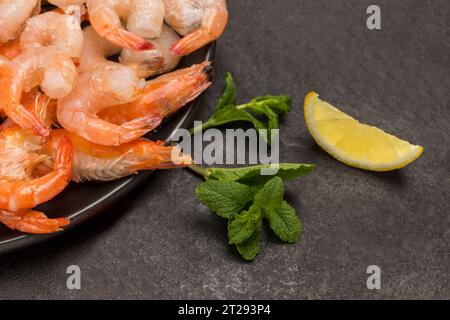 Tigergarnelen und Garnelen in schwarzer Schüssel. Tomaten, Knoblauchzehen, Zitrone auf dem Tisch. Schwarzer Hintergrund. Gesunde Ernährung. Flache Lagen Stockfoto