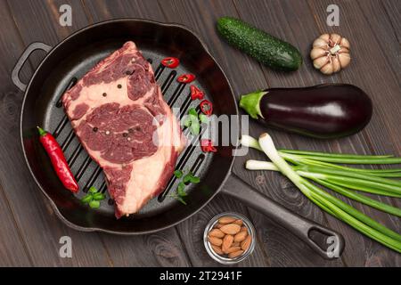 Rohes Fleisch. Marmoriertes Rindersteak in der Pfanne. Auberginen, Knoblauch und grüne Zwiebeln auf dem Tisch. Natürliche Proteinquelle. Flache Lagen. Dunkler Holzhintergrund Stockfoto