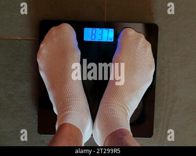 Füße mit weißen Socken auf Personenwaage, Körpergewicht messen, digitale Gewichtsanzeige in Kilogramm Stockfoto