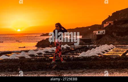 Eine junge Frau, die im orangefarbenen Sonnenuntergang in der salina spaziert und im Hintergrund den Leuchtturm von Fuencaliente auf der Route der Vulkane südlich der Stockfoto