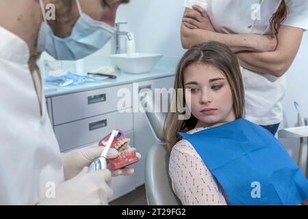 Junger Zahnarzt in weißer Uniform und medizinischer Schutzmaske mit Kiefermodell mit Zahnspangen für weibliche Patientin in der Zahnarztpraxis mit moderner Ausstattung. Stockfoto