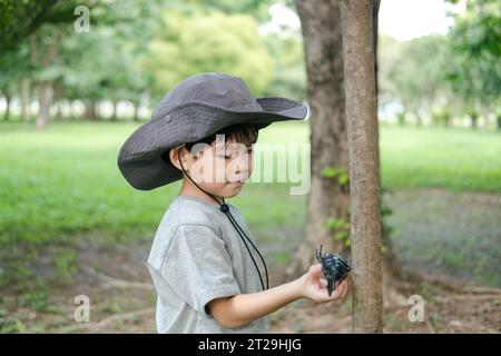 Ein asiatischer Junge, der einen Hut in einem Wald-Erkundungsanzug trägt, spielt mit einem Käfer an der Hand. Stockfoto