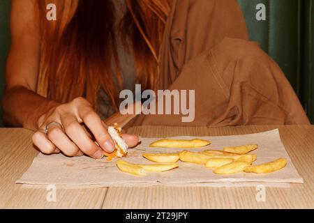 Schneiden Sie anonyme Frau mit roten Haaren am Tisch und genießen Sie Hamburger und Pommes Frites im Restaurant Stockfoto