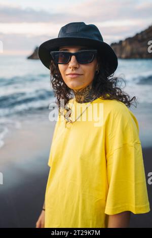 Porträt einer jungen Frau in lässiger Kleidung und Sonnenbrille, die auf sandigem Meer steht, während sie die Kamera vor dem wehenden Meer ansieht Stockfoto