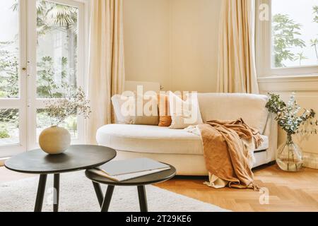 Wohnzimmer mit Kissen und Decke auf komfortabler Couch und Kaffeetischen, die durch Fenster in einem modernen Apartment platziert werden Stockfoto