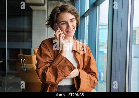 Glücklicher junger Büroangestellter in der Nähe des Fensters, der jemanden anruft, einen Anruf auf dem Mobiltelefon entgegennimmt, spricht und lächelt Stockfoto