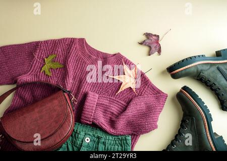 Flacher Pullover, grüne Jeans, Tasche, Stiefel und Herbstblätter auf neutralem Hintergrund. Herbstmodekonzept. Draufsicht, flach. Stockfoto