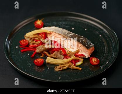 Gegrilltes Lachssteak auf einem Holzbrett mit einem frischen gesunden Salat, Zitrone, Gemüse, Kräutern und Gewürzen auf einem dunklen Stockfoto