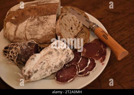 Wurst-Wurst und handwerkliches Sauerteigbrot von den lokalen Märkten, geschnitten mit dem geöffneten Opinel-Messer auf einem weißen Teller auf einem Eichentisch Stockfoto