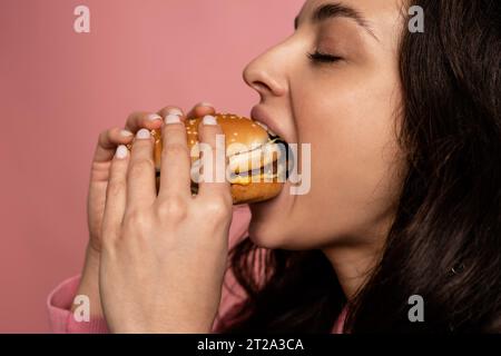 Süße junge Frau, die während des Fotoshootings in einem Studio mit geschlossenen Augen an einem Sandwich knabbert. Hunger und Lieblings-Fast-Food-Konzept Stockfoto