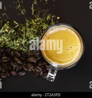 Eine frisch gebrühte Tasse Kaffee in einer Kaffeetasse aus Glas, neben Kaffeebohnen und Teeblättern auf schwarzem Hintergrund. Stockfoto