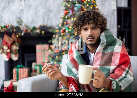 Porträt des kranken Mannes zu Hause auf Sofa, Mann feiert Neujahr und weihnachten, sitzt am Wintertag im Wohnzimmer Nahaufnahme und Blick auf die Kamera, in der Nähe dekorierter Weihnachtsbaum, Grippe und Kälte. Stockfoto