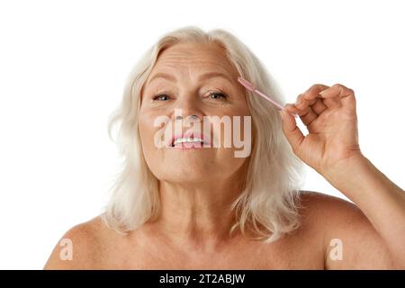 Elegante, schöne Seniorin, die sich um die Brauen kümmert und mit einem Pinsel vor weißem Studiohintergrund putzt Stockfoto