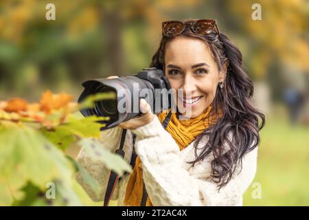 Schöne Frau, die eine professionelle Kamera hält und nach Komposition in herbstlicher Natur sucht, lächelt sie. Stockfoto