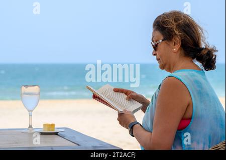 Das Leben ist ein Strand: Eine indische Frau mittleren Alters sitzt bei einem Drink und liest ein Buch mit Strand und Meer als Hintergrund. Mui Ne, Vietnam. Stockfoto