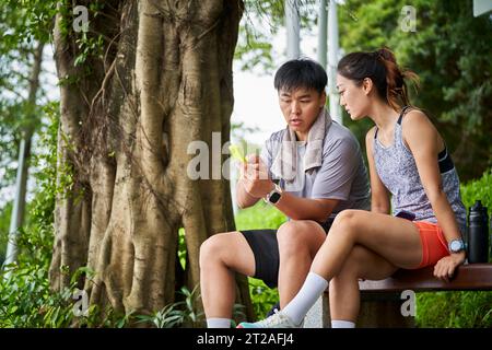 Junges asiatisches Paar, das auf das Handy schaut, während er eine Pause während des Outdoor-Trainings macht Stockfoto