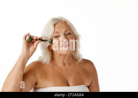 Schöne ältere Frau in ihren 60ern, mit faltigem Gesicht, das Gesichtsmassage mit Spezialwerkzeug vor weißem Studiohintergrund macht Stockfoto